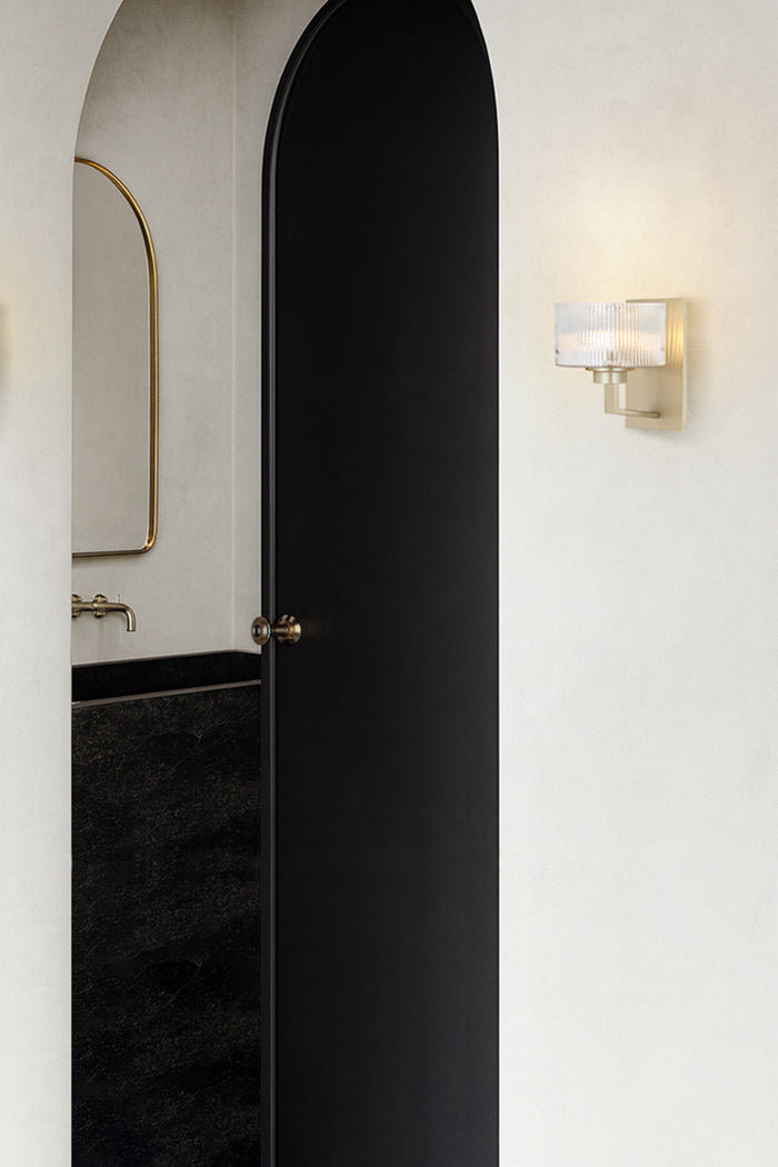 Antique Silver wall sconce with a reeded glass shade as hall lighting next to an arched doorway od a white, black and gold bathroom space.