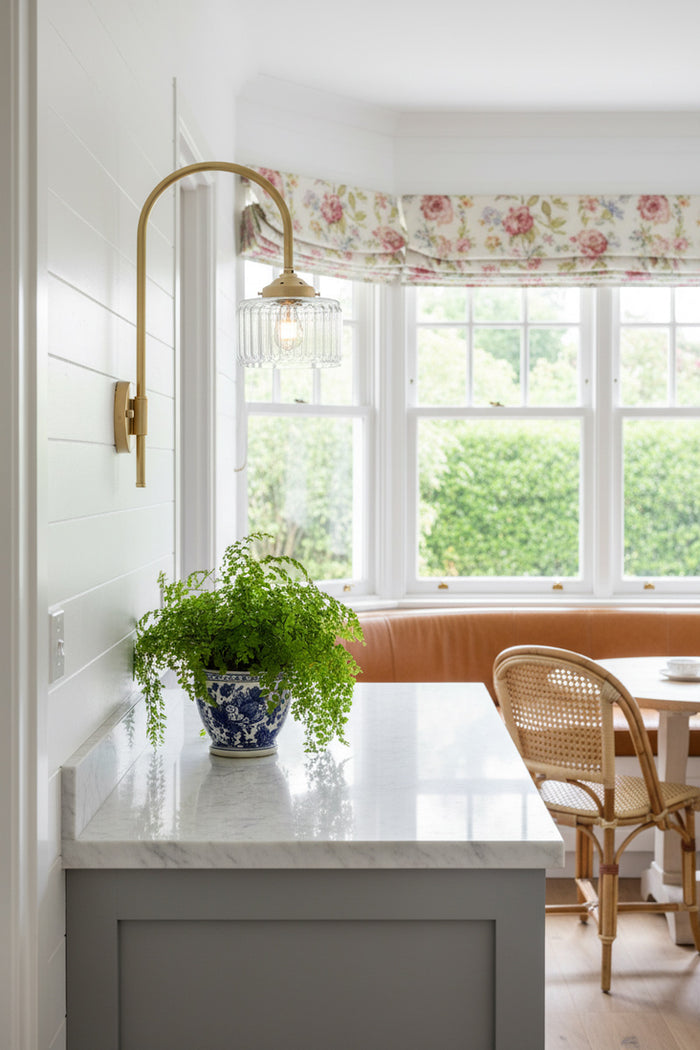 Gold/brass shepherds hook wall light with a clear glass shade, over a side bench in a bright breakfast nook.