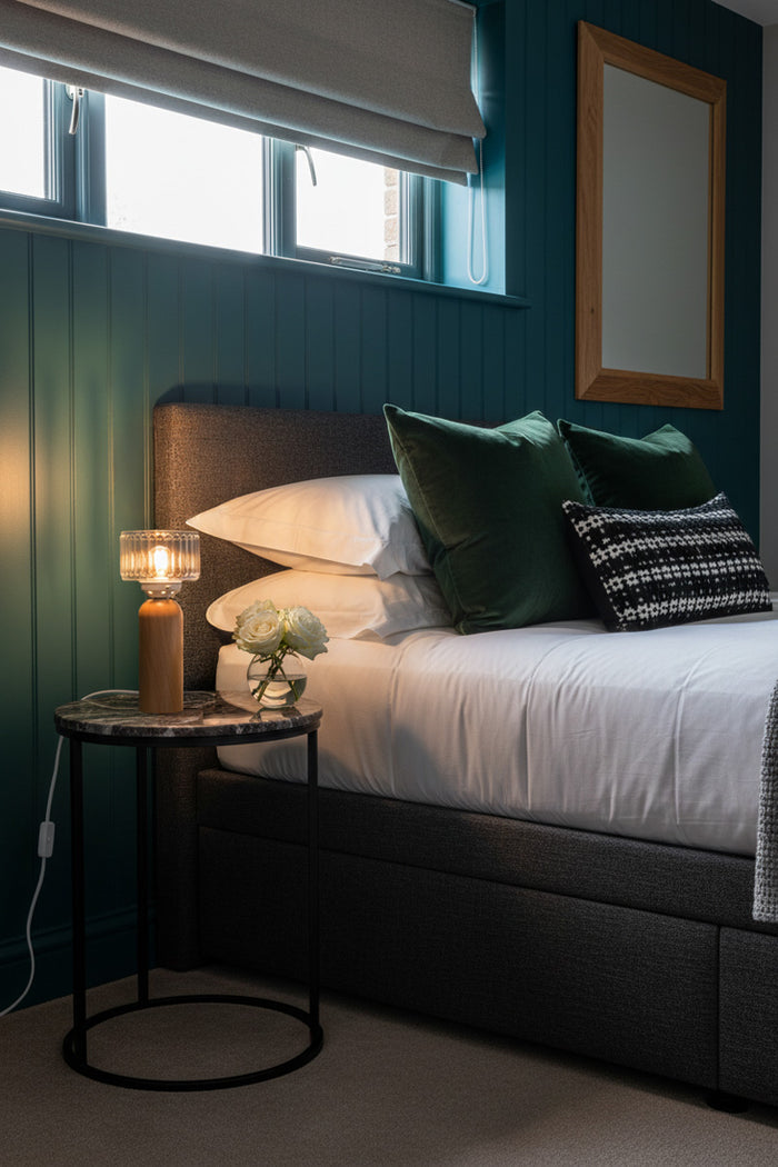 bedroom featuring a bed with white bedding, dark green pillows, and a black upholstered headboard, set against a dark teal paneled wall. A lit wooden glass table lamp sits on a small marble-top side table next to the bed.