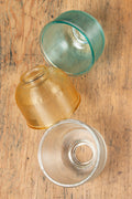 Three small holophane style glass shades in blue, clear and amber, sitting on a timber bench at varying angels.
