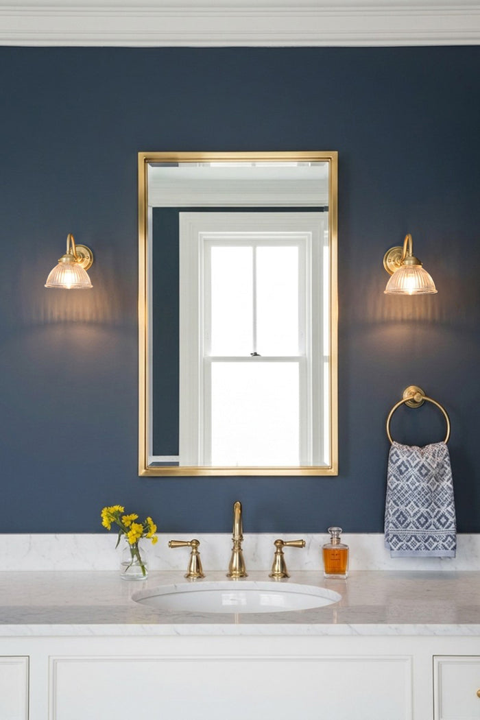 A framed wall mirror with a gold frame hangs on a dark navy blue wall above a white marble bathroom vanity. Two matching gold brass gooseneck wall lights with ribed glass light the space.