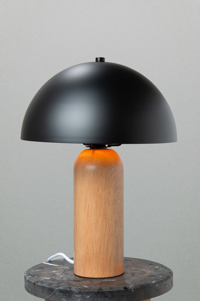 Table lamp with a black metal dome shade and rounded wood base with a natural finish. Lamp placed on a marble look side table.