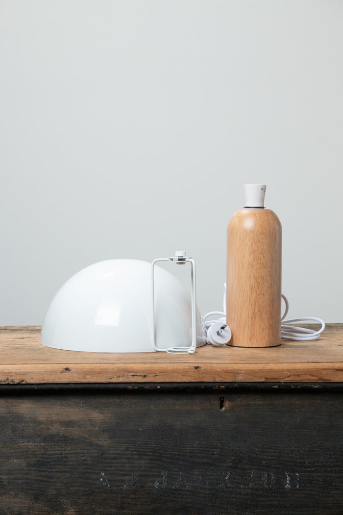 Table lamp with a white metal dome shade and rounded wood base with a natural finish, uninstalled on a timber bench.