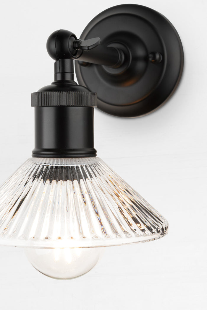 Petite pleated clear glass shade with clear bulb on a short, black wall sconce.