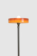 Front view detail of floor lamp showing circular amber shade with exposed globe and slim dark stem.
