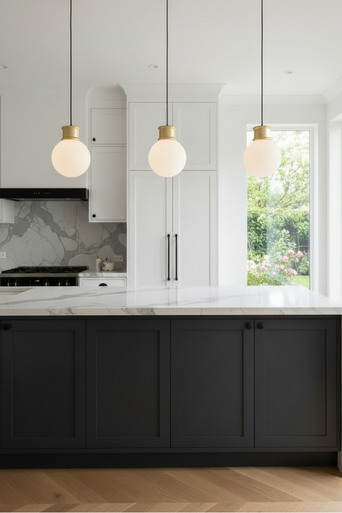 Modern kitchen interior with three pendant lights featuring white opal large shades and brushed brass fixtures, hanging above a dark charcoal island with a white marble countertop.