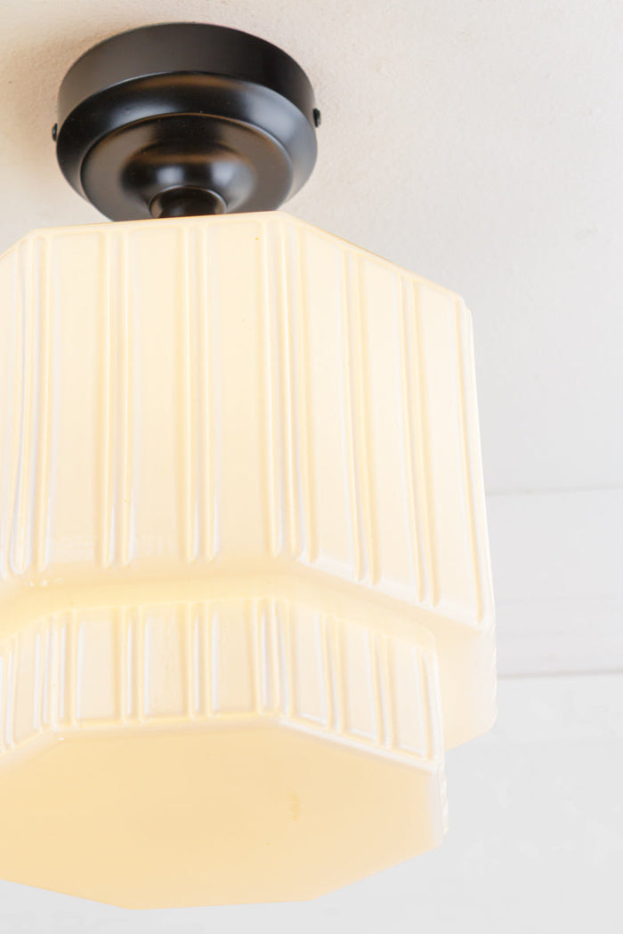 Flush mount light consisting of an opal glass shade with step silluette and unique fluted detailing, paired with a black batten holder and gallery.