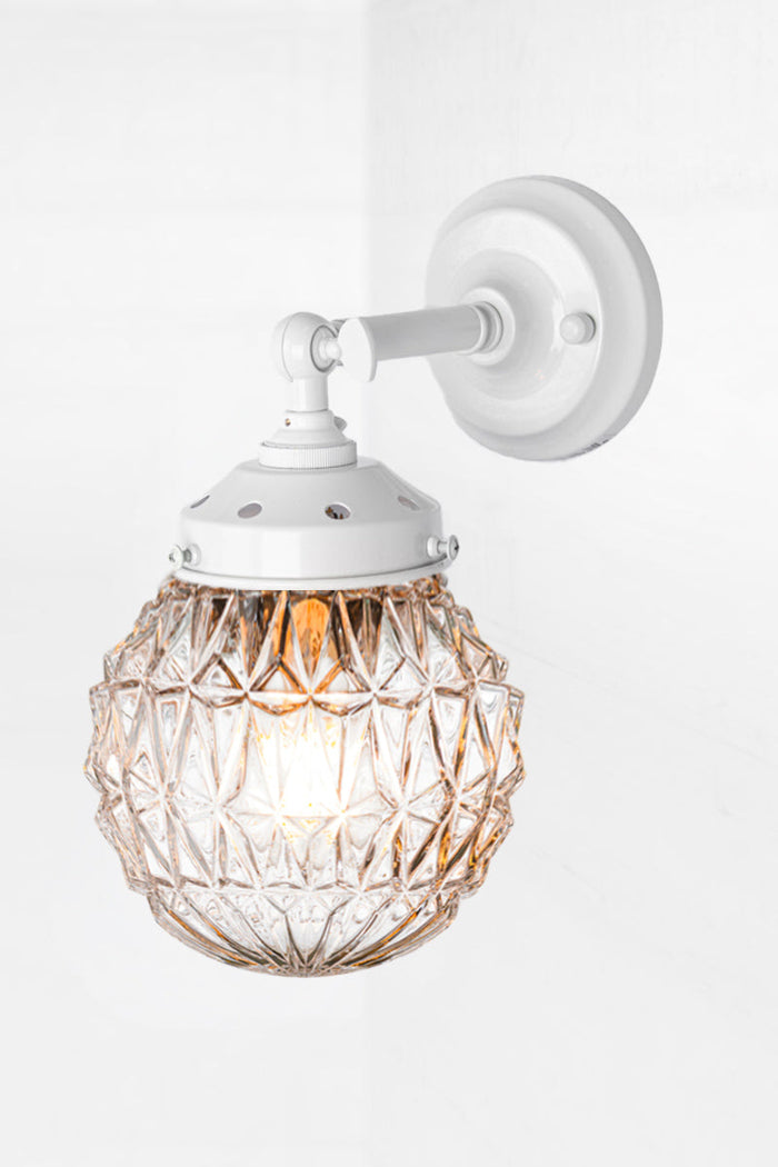 Short white, straight arm wall sconce with tiltable lampholder, paired with a clear glass orb shade with detailing.