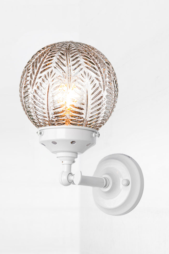 Short white, straight arm wall sconce with tiltable lampholder, paired with a clear glass orb shade with intricate detailing.