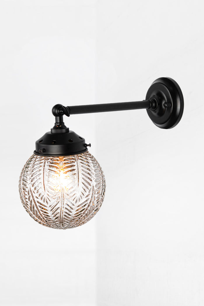 Long straight arm wall sconce with tiltable lampholder in black paired with a clear glass orb shade with intricate detailing.