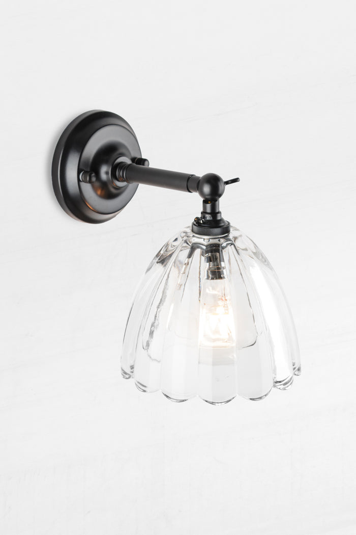 Black short straight arm wall sconce with a clear glass shade with a scalloped edge.