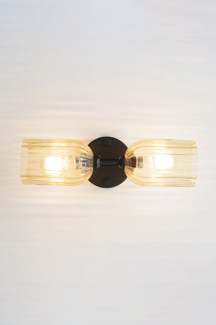 Black double wall sconce with two elongated amber glass shades with fluted detailing installed horizontally.