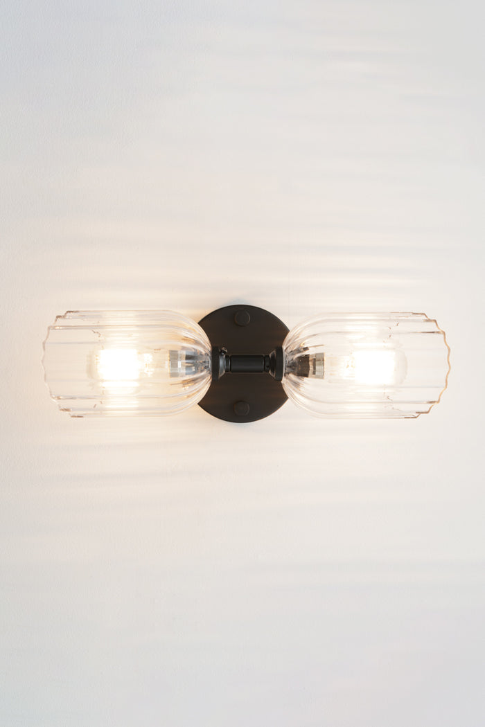 Black double wall sconce with two elongated clear glass shades with fluted detailing installed horizontally.