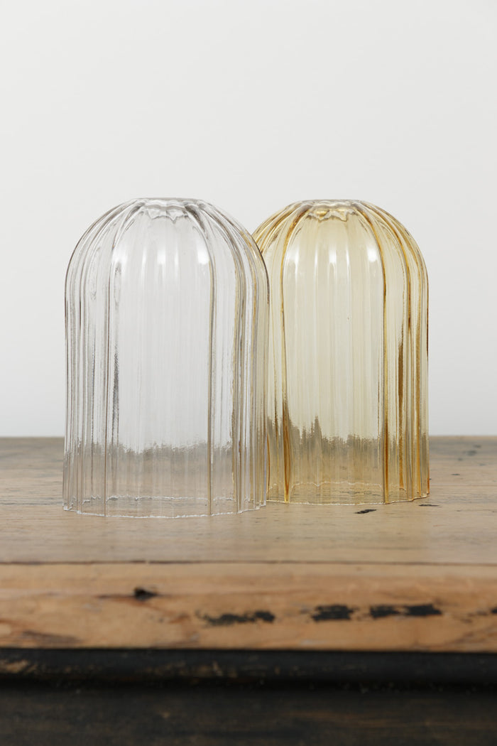 Two glass shades with fluted detailing in amber or clear with an angled cut edge, placed on a timber bench.