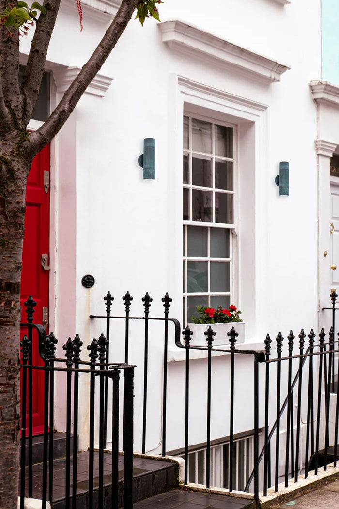 Two up/down brass wall spot lights with a green patina finish on a white front facade with a red front door.