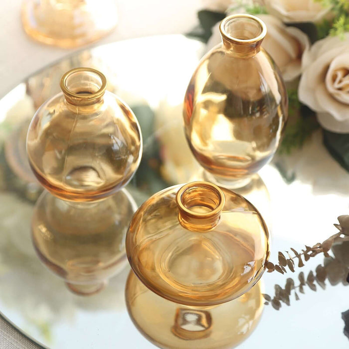 Three amber glass vases on a reflective surface with floral accents.